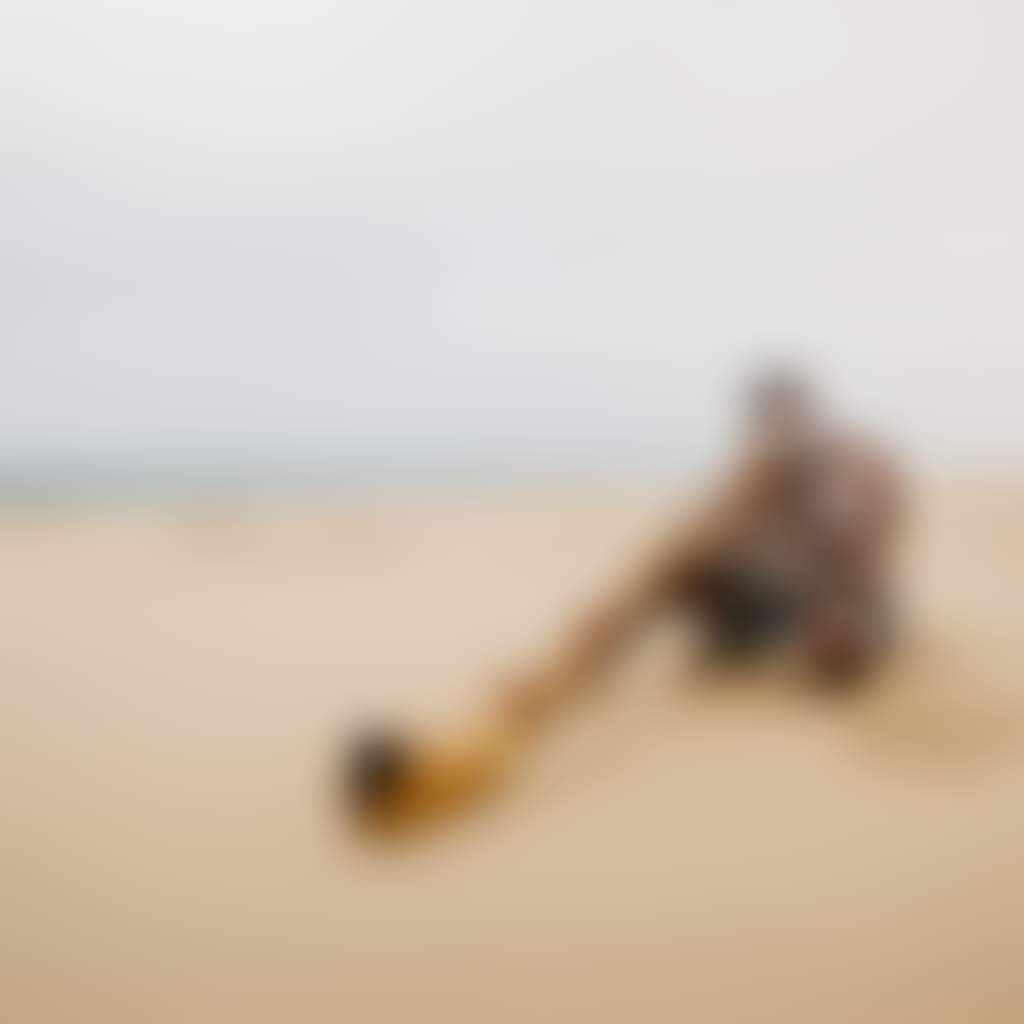 A person is sitting cross legged on the beach and playing didgeridoo
