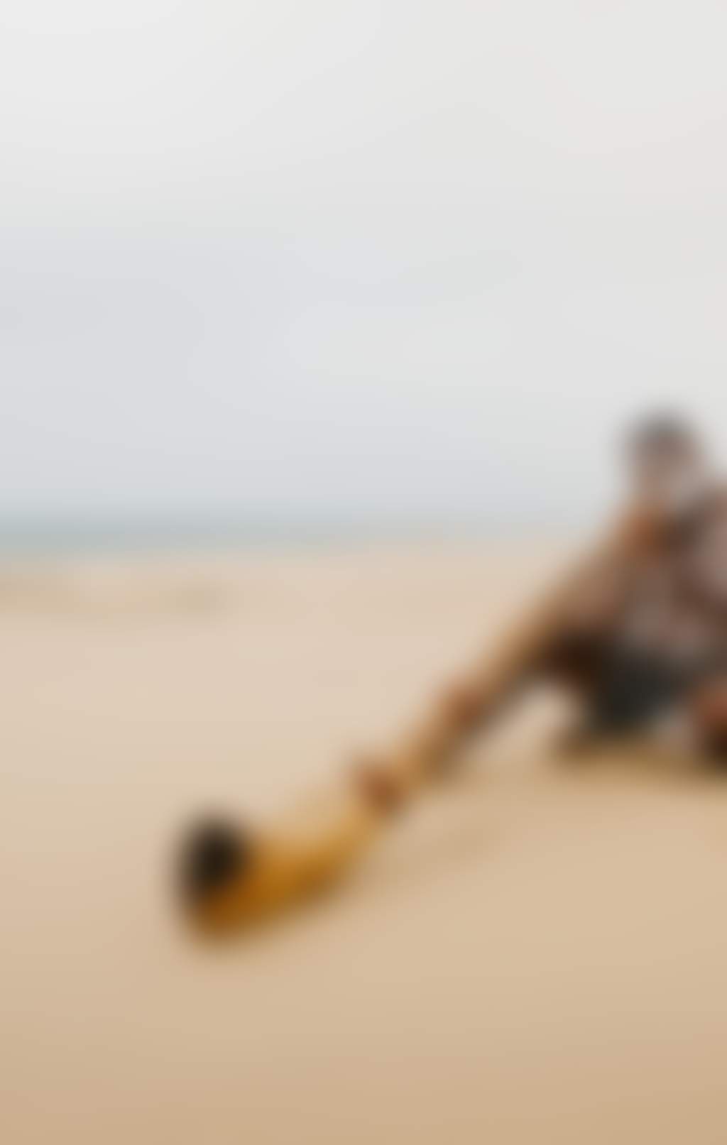 A person is sitting cross legged on the beach and playing didgeridoo