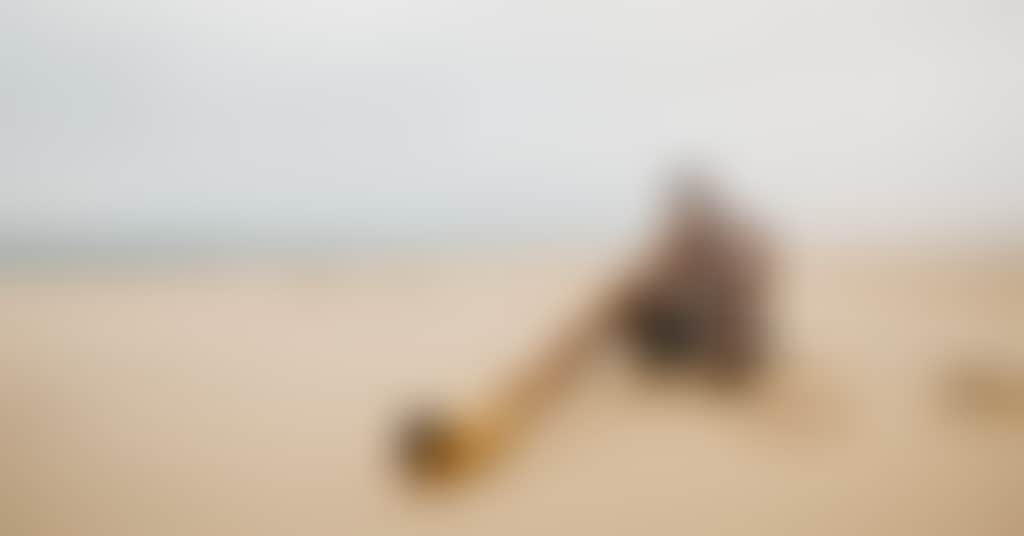 A person is sitting cross legged on the beach and playing didgeridoo