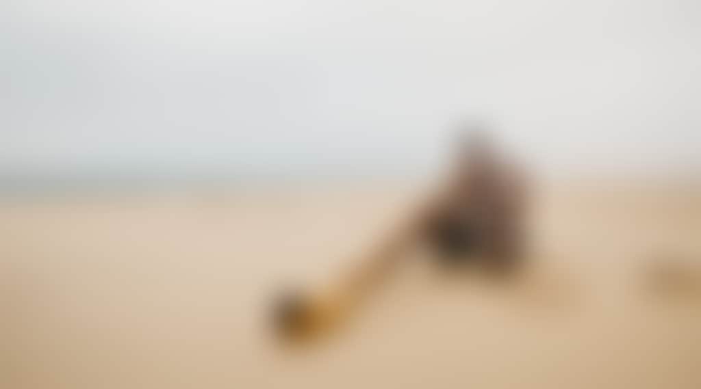 A person is sitting cross legged on the beach and playing didgeridoo